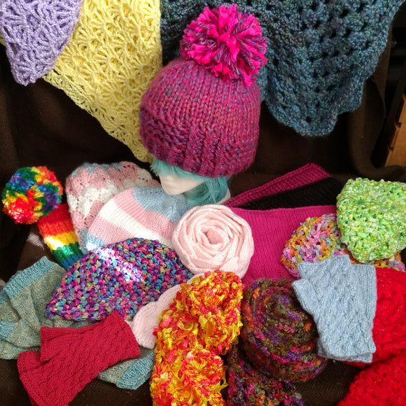A display of numerous assorted knitted and crocheted accessories; hats, scarves, gloves, shawls, and more, with a pink hat with a giant pompom modeled on a styrofoam mannequin head.