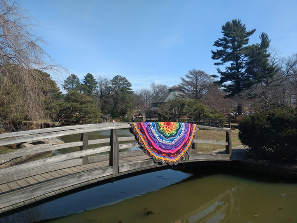 A landscape image of a curved footbridge over a small body of water. Various types of trees are visible in the background, and a colorful crochet round blanket is displayed on the bridge handrail.